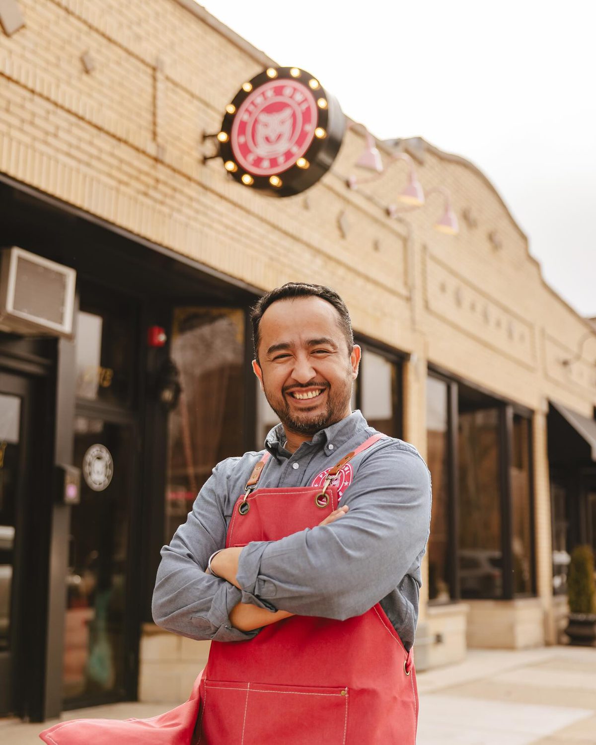 Alex Herrera in a red apron, arms crossed, standing on the sidewalk under the lit Pink Owl sign