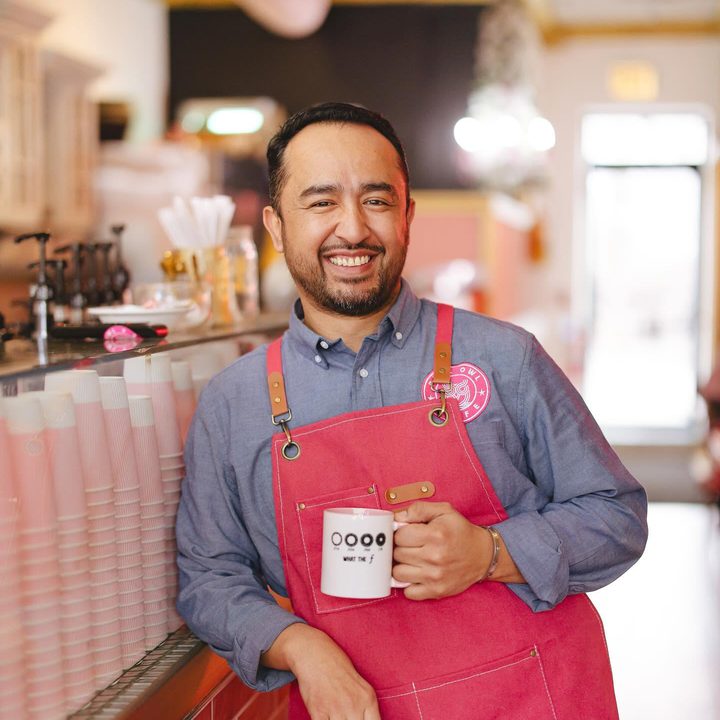Alex Herrera in a red apron, holding a mug at the counter, smiling