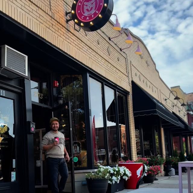 Alex stepping out of the cafe doorway holding a pink cup, under the lit marquee sign